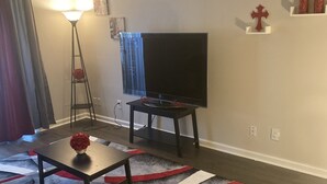 Un salon avec un téléviseur à écran plat posé sur un meuble, une lampe sur pied, une petite table avec un vase et un tapis à motifs.