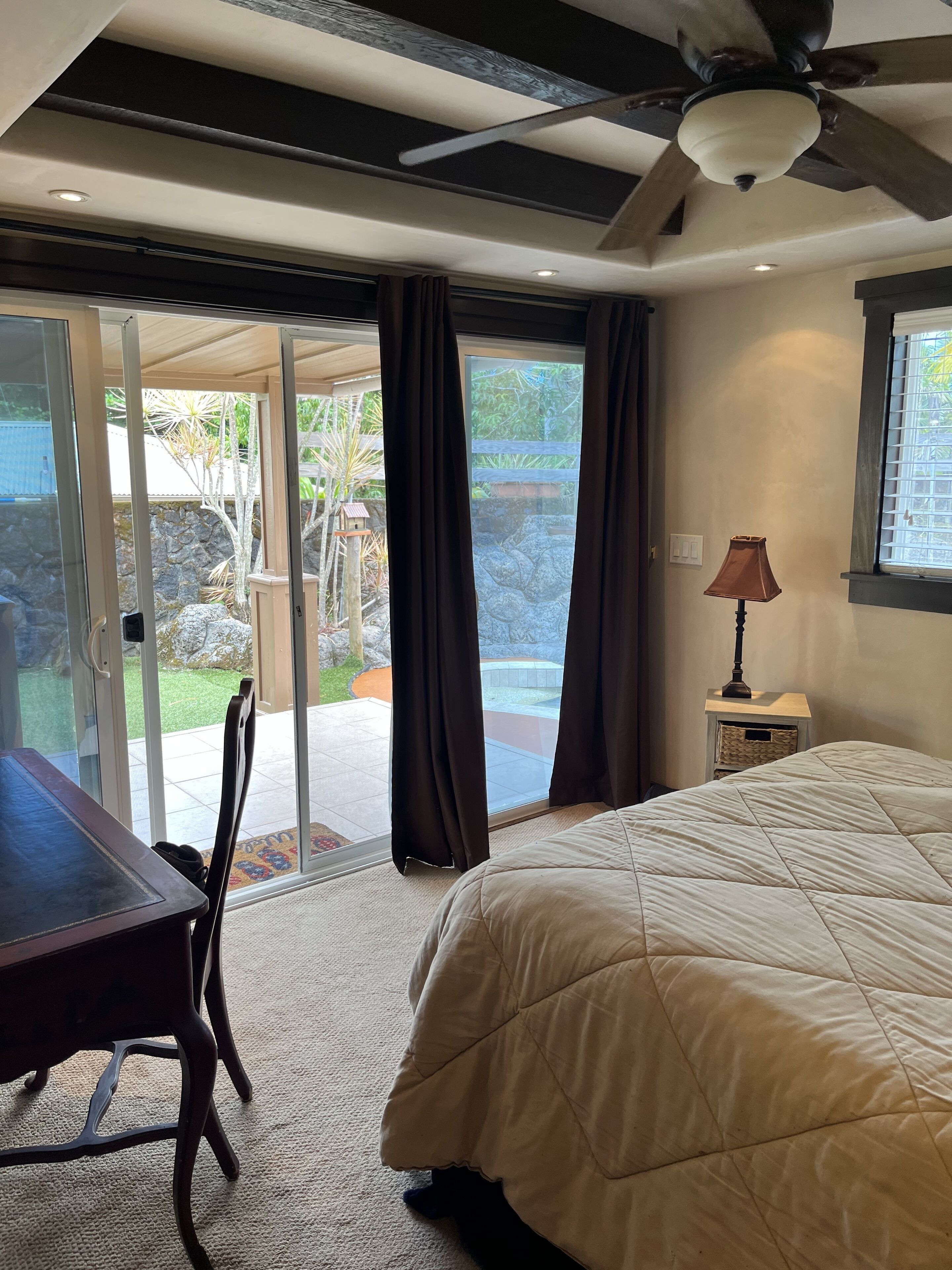 A bedroom with a bed, a desk, and a chair. There is a window with curtains, a ceiling fan, and a lamp on a nightstand.
