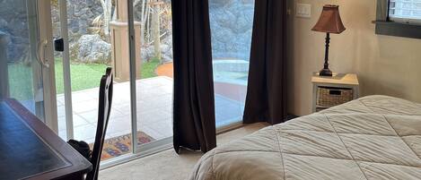 A bedroom with a bed, a desk, and a chair. There is a window with curtains, a ceiling fan, and a lamp on a nightstand.