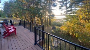 A wooden deck with red Adirondack chairs, a table, and a view of a forest and lake.