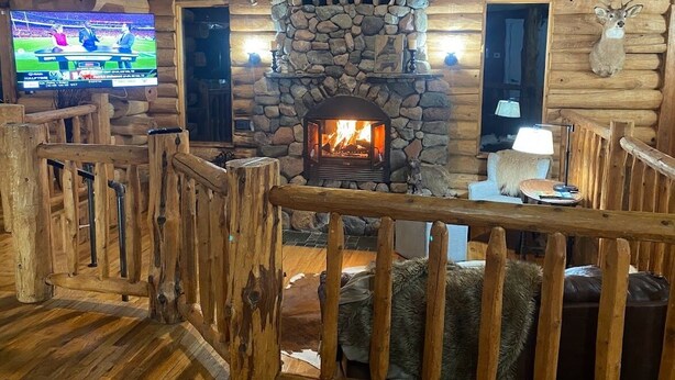 A log cabin interior with a stone fireplace, wooden walls, and a mounted deer head.