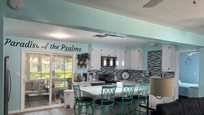 A modern kitchen with teal and white cabinetry, a central island with bar stools, and a dining area with a black sofa. - Weeki Wachee Gardens on the Canal (Spring Hill)