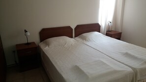 Uma cama de casal com cabeceira de madeira, uma mesa de cabeceira com abajur e uma mesa lateral com luminária.