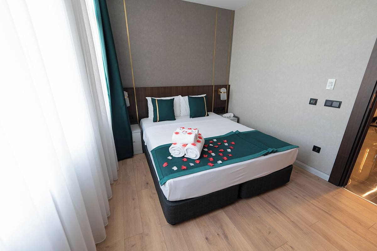 Ein Hotelzimmer mit einem großen Bett, einem hölzernen Kopfteil, einer grünen Decke und einem ordentlich bezogenen Bett mit Handtuch und Rosenblüten.