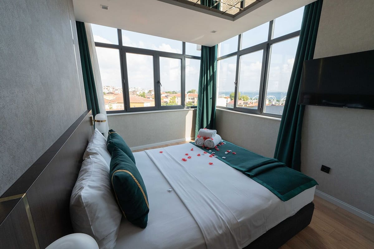 Ein Hotelzimmer mit einem großen Bett, Blick auf die Stadt durch große Fenster und einem an der Wand montierten Flachbildfernseher.