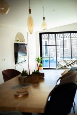 A dining area with a wooden table, chairs, hanging pendant lights, a potted plant, and a view of a pool through large windows.