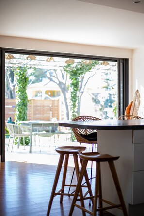 A modern kitchen with a large window, wooden stools, and a wicker chair.