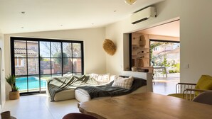 Ein modernes Wohnzimmer mit einem großen Schiebeglasfenster, das zu einem Pool führt, einem Holzessttisch und einer gemütlichen Sitzecke.