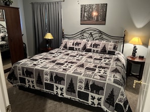 Quarto com uma cama, duas mesas de cabeceira, um criado-mudo, uma luminária, um quadro na parede e uma porta.