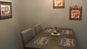 Una mesa de comedor con acabado en madera oscura, rodeada por cuatro sillas. Sobre la mesa, hay un candelabro con múltiples luces. En las paredes, dos cuadros con la palabra “Apple” y un adorno mural. Debajo de la mesa, hay estantes con diversos objetos.