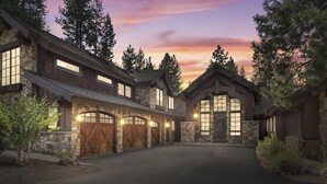 A large, stone-clad house with multiple garage doors and large windows, surrounded by trees and a driveway. - Luxury Sunriver Estate | 5 Kings + Bunk Rm | Huge Spa & Arcade | Chef's Kitchen (Sunriver)