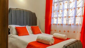 Ein ordentlich bezogenes Bett mit weißer und roter Bettwäsche, einem grauen Kopfteil und einem Fenster mit gemusterten Vorhängen.