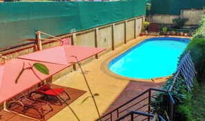 Una piscina de borde curvo, rodeada por una terraza de madera y una valla verde. Hay una sombrilla roja y una silla metálica cerca de la piscina.