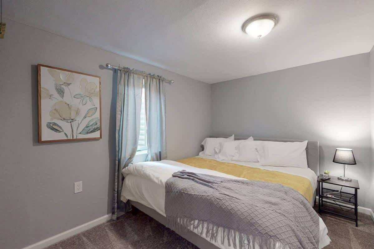 A bedroom with a bed, a nightstand, a lamp, a window with curtains, and a framed picture on the wall.