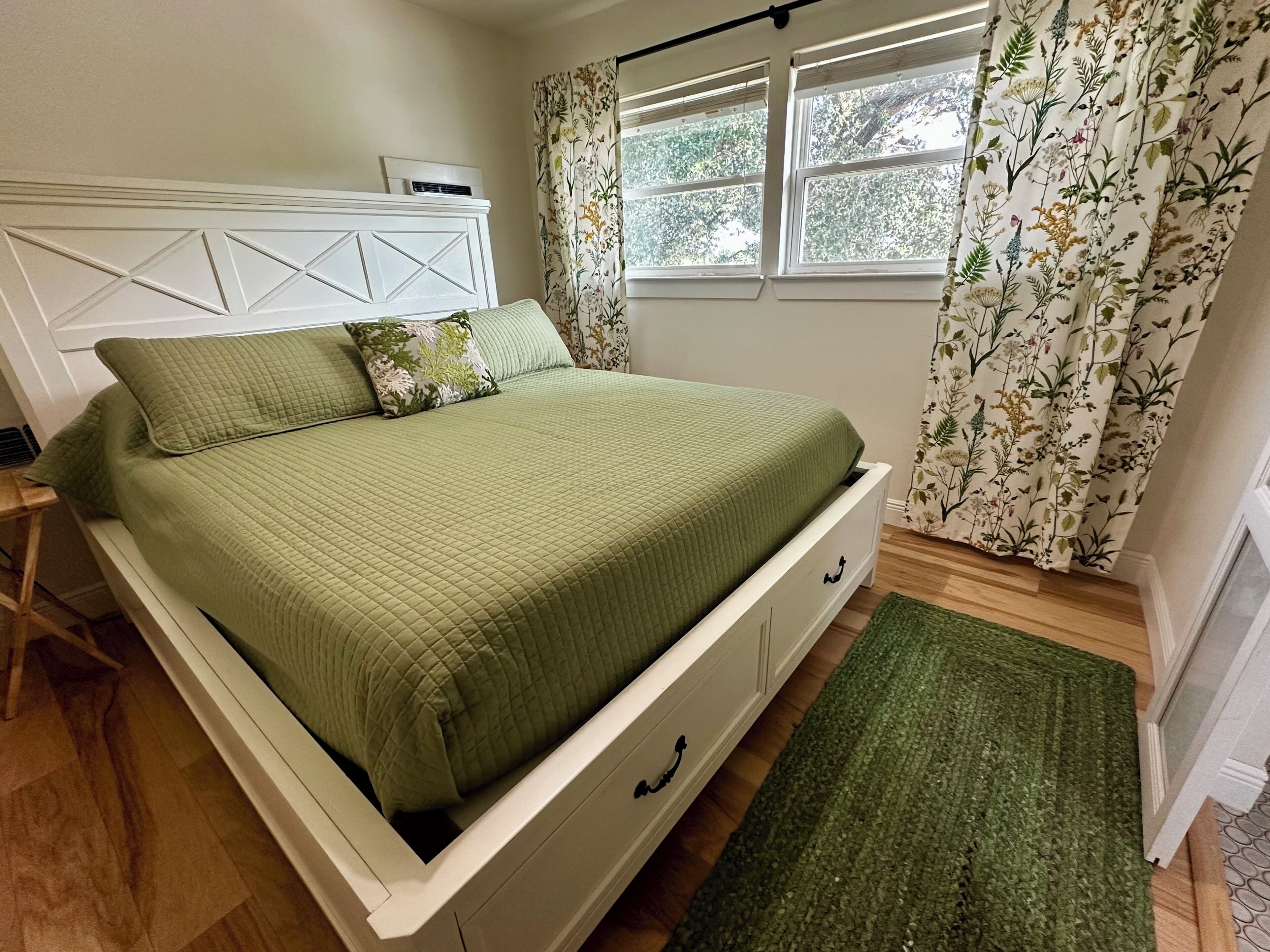 Een netjes opgemaakt bed met een groene quilt, een wit hoofdbord en een raam met bloemen gordijnen.