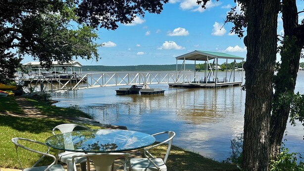 A lakeside patio with a glass table and metal chairs, a covered seating area, and a dock extending into the water. - Waterfront Property on PK - 7 miles away from Rocker B baseball facility  (Graford)