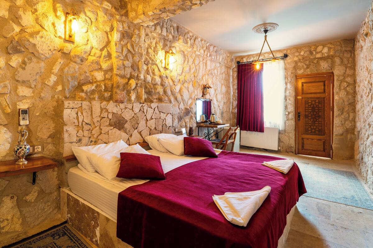 Un dormitorio con paredes de piedra, una cama, una mesita de noche, una ventana con cortinas y una puerta de madera.
