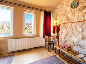 Una habitación con pared de piedra, ventana con vistas a formaciones rocosas, radiador, una silla de madera y estante con toallas.