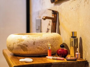 A bathroom vanity with a large, round, beige sink, a gold faucet, and various toiletries including a perfume bottle, a compact, and a small bottle. - Ala Stone Hotel (Nevsehir)