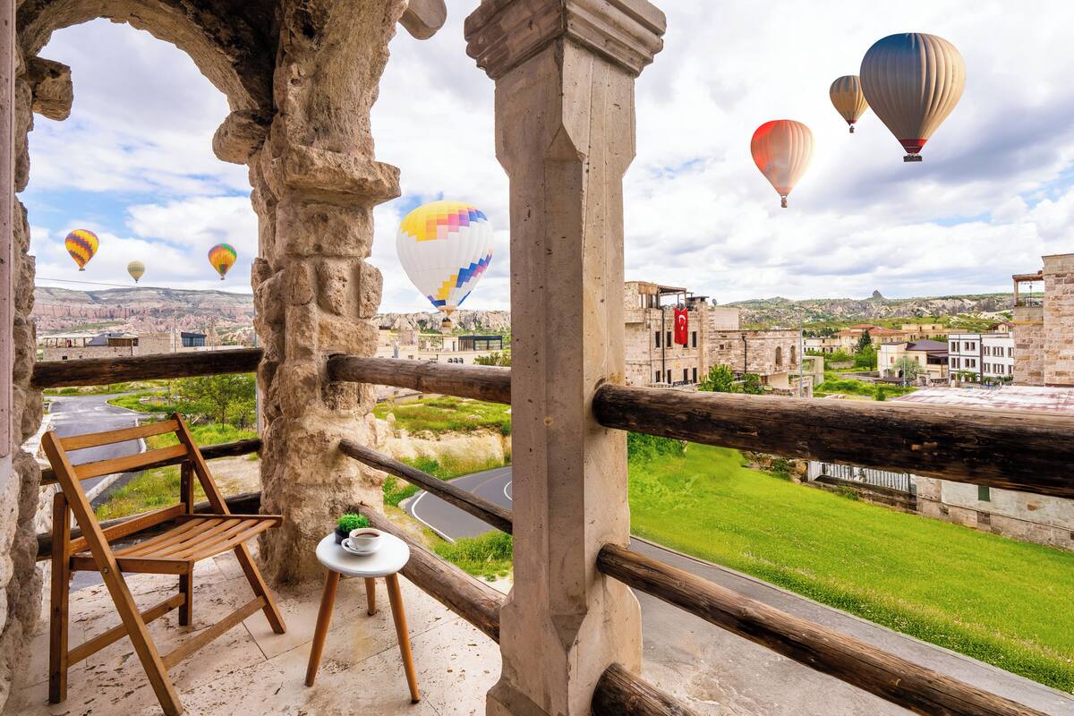 Un balcón con una silla de madera, una mesa pequeña con una taza, y una vista de un pueblo con edificios y globos aerostáticos a lo lejos.