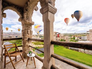 A balcony with a wooden chair, a small table with a cup, and a view of a town with buildings and hot air balloons in the distance. - Ala Stone Hotel (Nevsehir)