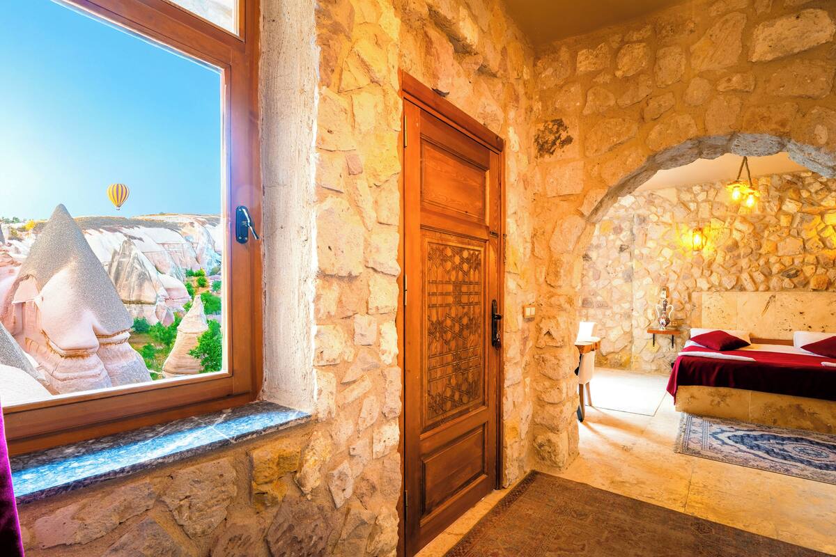 Una habitación con paredes de piedra, una puerta de madera, una cama con ropa de cama roja, una mesita con una lámpara y vistas a un paisaje con formaciones rocosas y un globo aerostático.