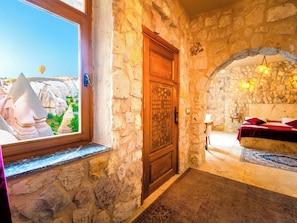 Una habitación con paredes de piedra, una puerta de madera, una cama con ropa de cama roja, una mesita con una lámpara y vistas a un paisaje con formaciones rocosas y un globo aerostático.