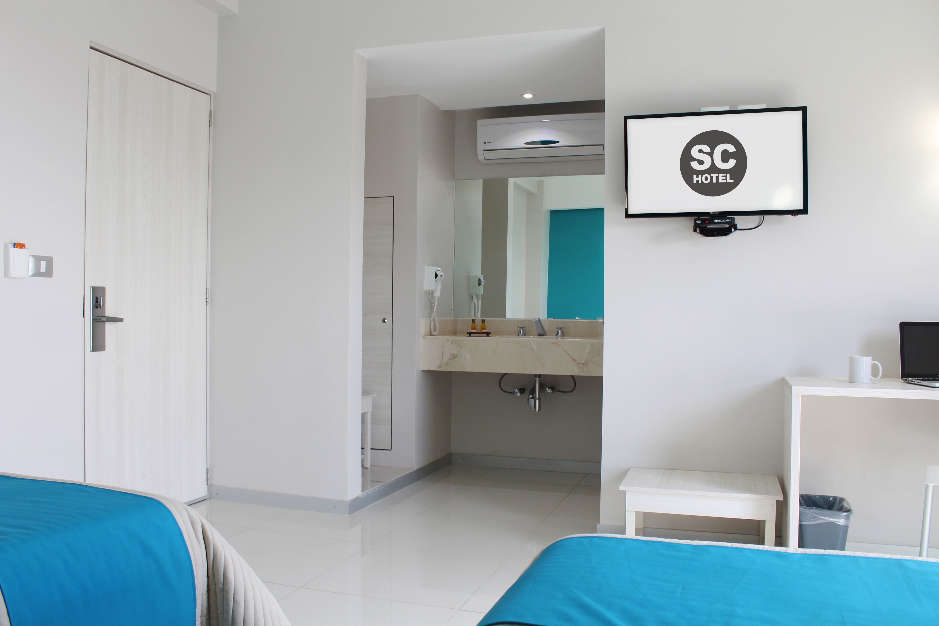 A hotel room with a bed, a desk, a TV, a bathroom with a mirror, and an air conditioning unit.