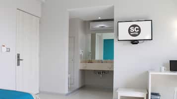 A hotel room with a bed, a desk, a TV, a bathroom with a mirror, and an air conditioning unit.