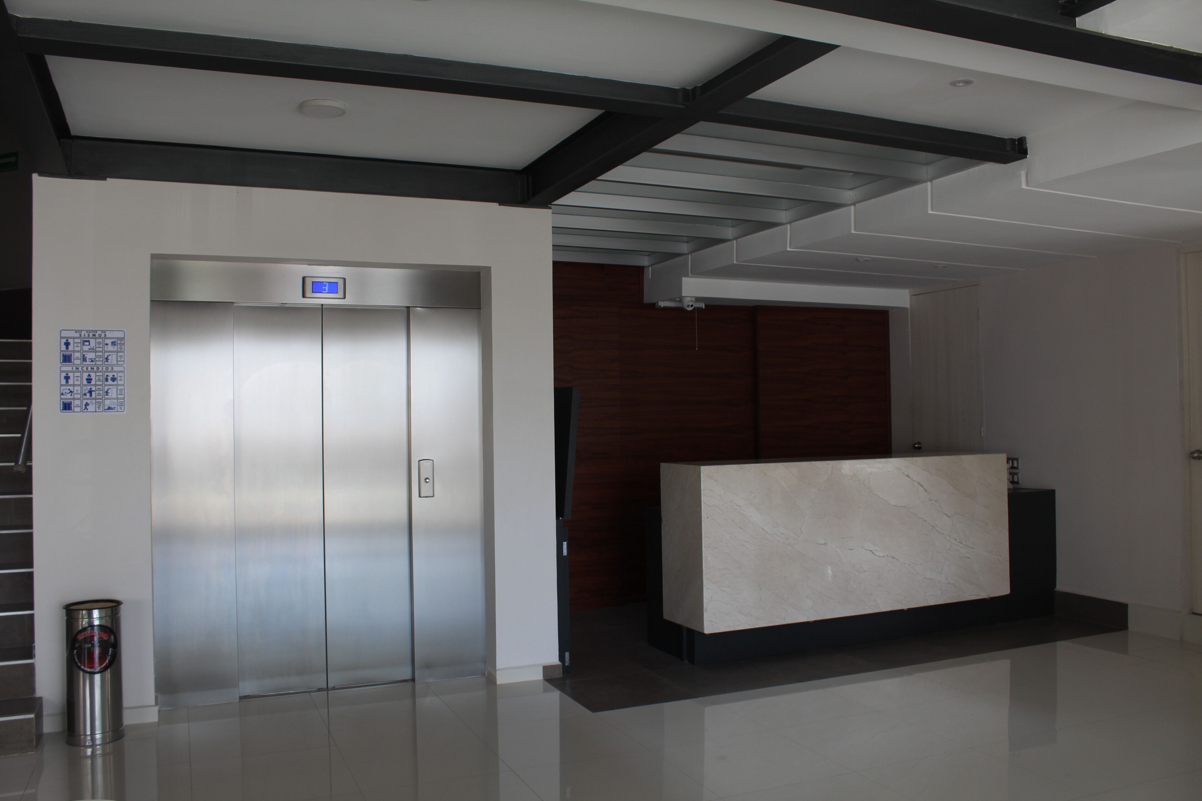 A modern reception area with a marble reception desk, a digital elevator display, and a trash bin.