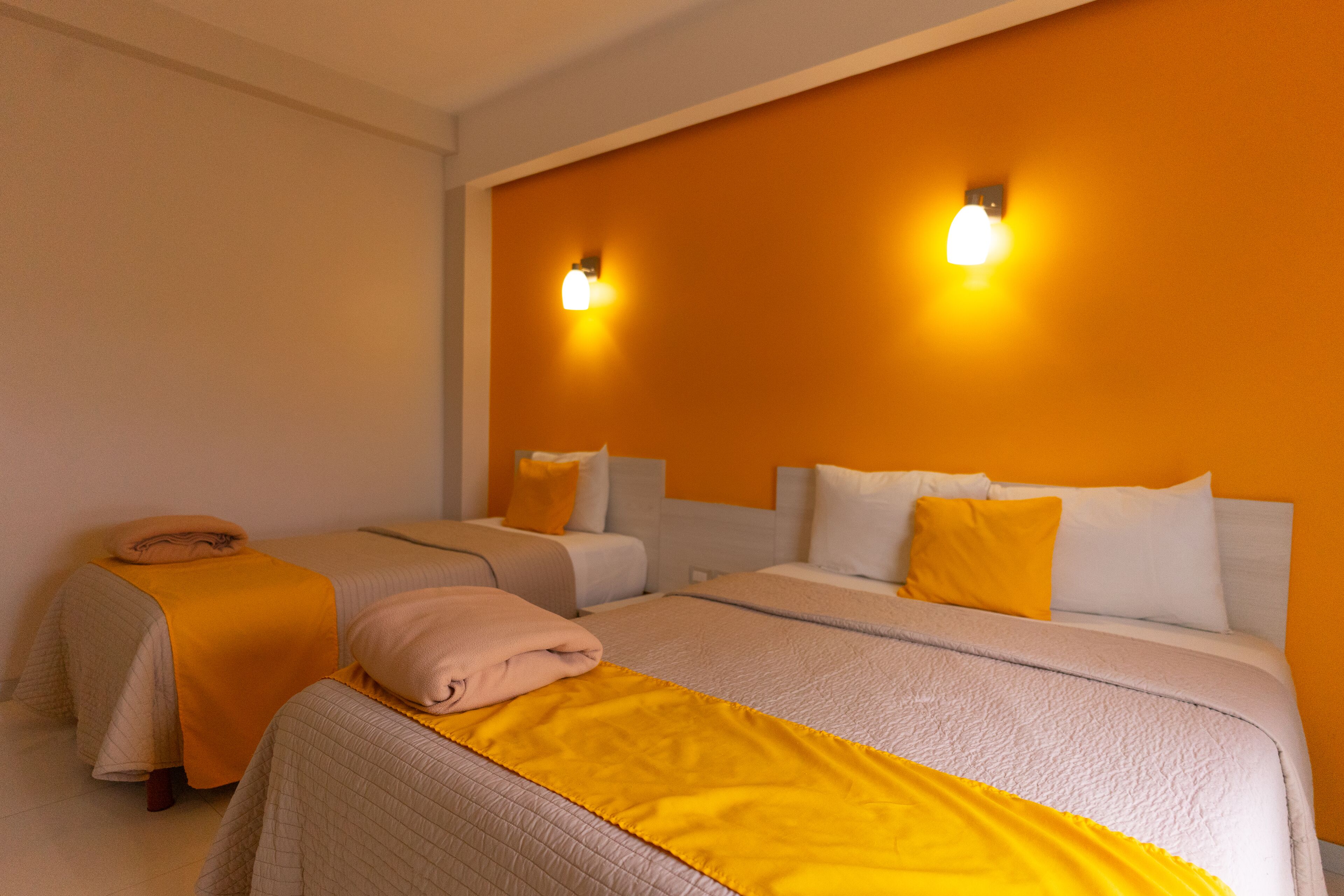 Habitación de hotel con dos camas, cada una con una manta y almohadas amarillas, y dos luces fijadas a la pared.
