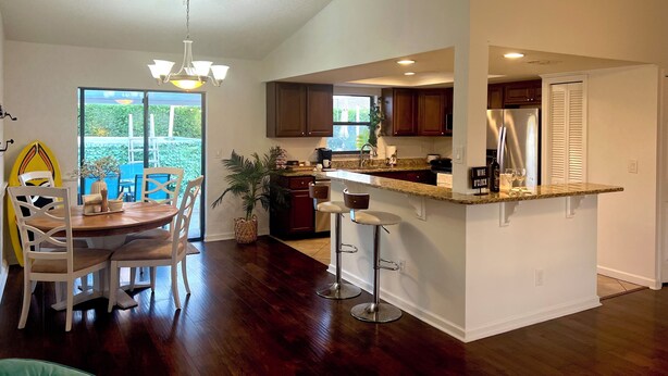 A modern kitchen with dark wood flooring, a central island with bar stools, and a dining area with a wooden table and chairs. - The Sunrise House (Port Richey)