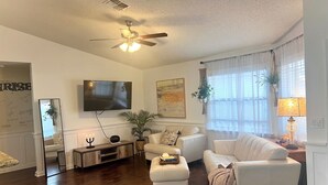A living room with a ceiling fan, a flat-screen TV, a large mirror, and two white leather sofas. - The Sunrise House (Port Richey)