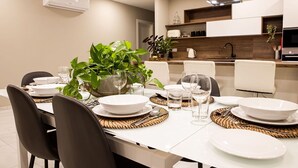 A modern dining area with a white table set for four, surrounded by black chairs. There is a potted plant on the table, and the room has a kitchen in the background with white cabinets and a wooden countertop.
