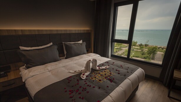 A neatly made bed with rose petals and a towel sculpture, a large window with a view of the sea, and a modern headboard. - Gursoy Exclusive Hotel (Arsin)