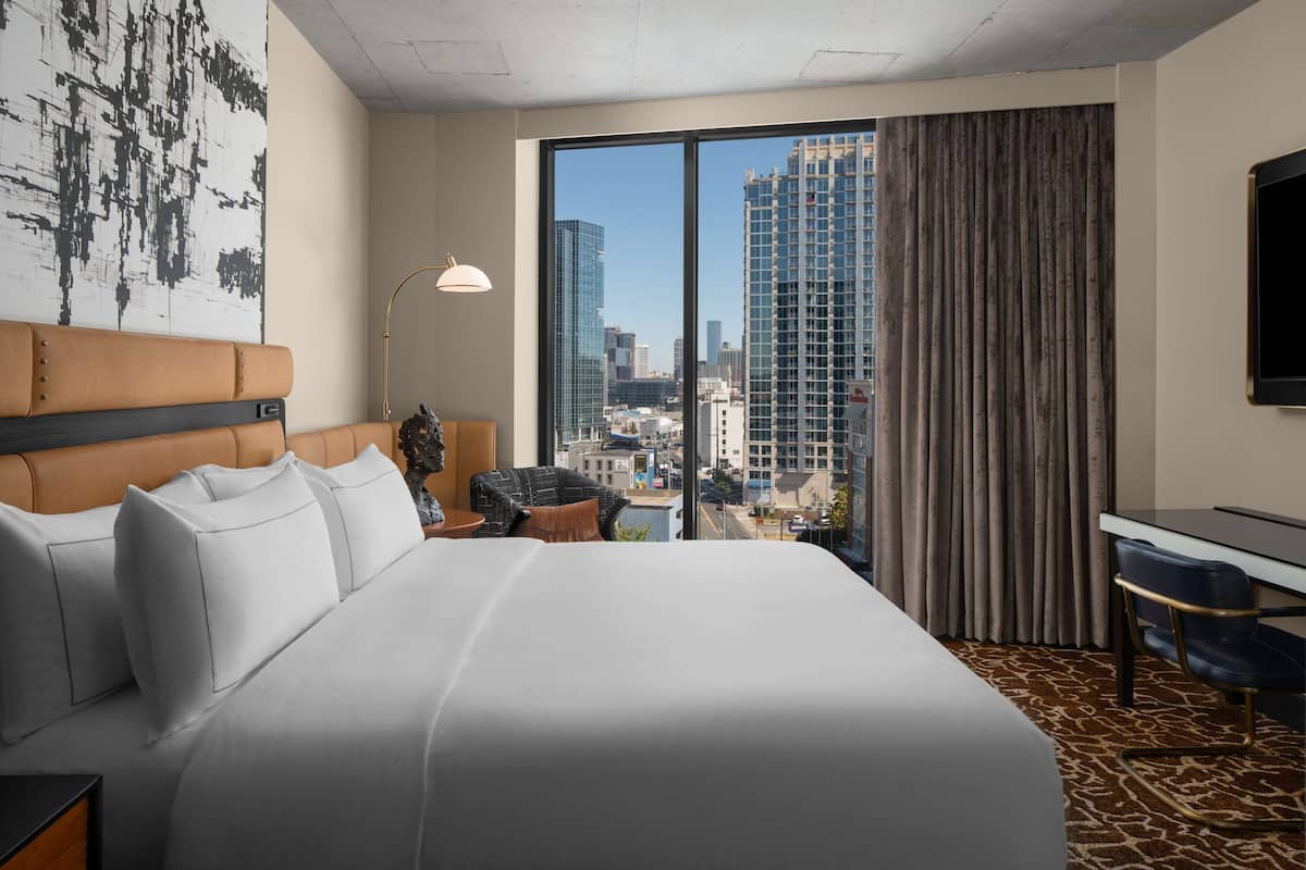 Een hotelkamer met een groot bed, een bureau, een stoel, een lamp en een uitzicht op de skyline door grote ramen.