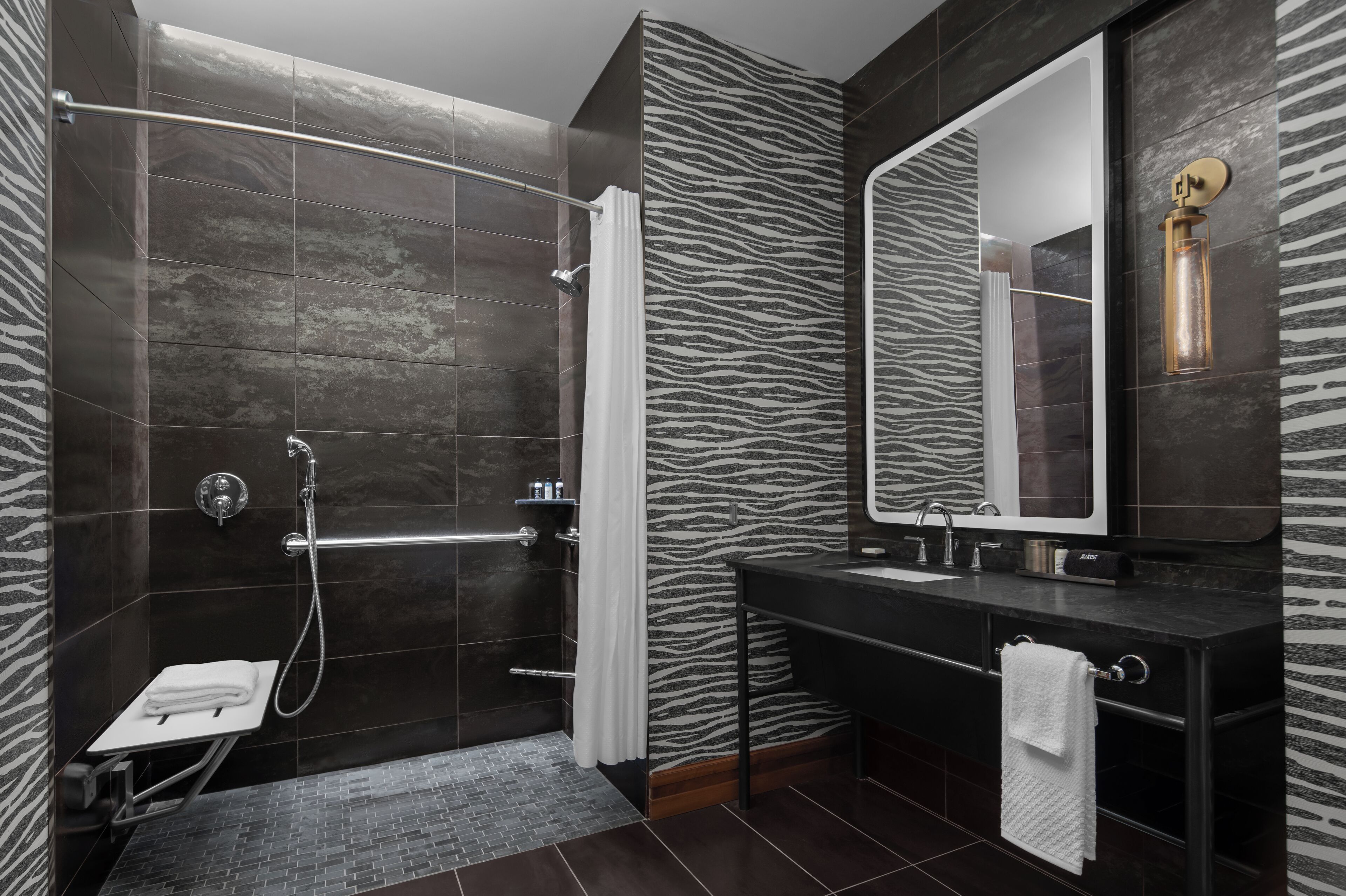 Une salle de bain moderne avec un mur à motifs zébrés, une douche en verre, un lavabo noir et un miroir doté d’un luminaire.