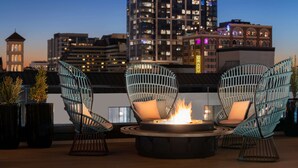 Een dak terras met twee rieten stoelen, een ronde tafel met vuurkorf, en een stadsachtergrond op de achtergrond.
