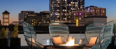 Una terraza en la azotea con dos sillones de mimbre, una mesa redonda con una chimenea y un horizonte urbano de fondo.