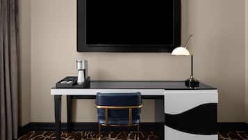 A hotel room with a desk, a chair, a TV mounted on the wall, a lamp, and a coffee machine.