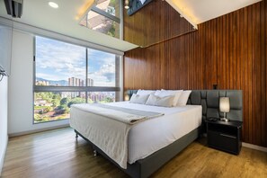 Relax in a spacious bedroom with a large bed against a wall of vertical slats, featuring a city view and a bedside table with a lamp.