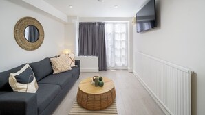 A modern living room with a grey sofa, a round wooden coffee table, a woven mirror, and a flat-screen TV mounted on the wall.