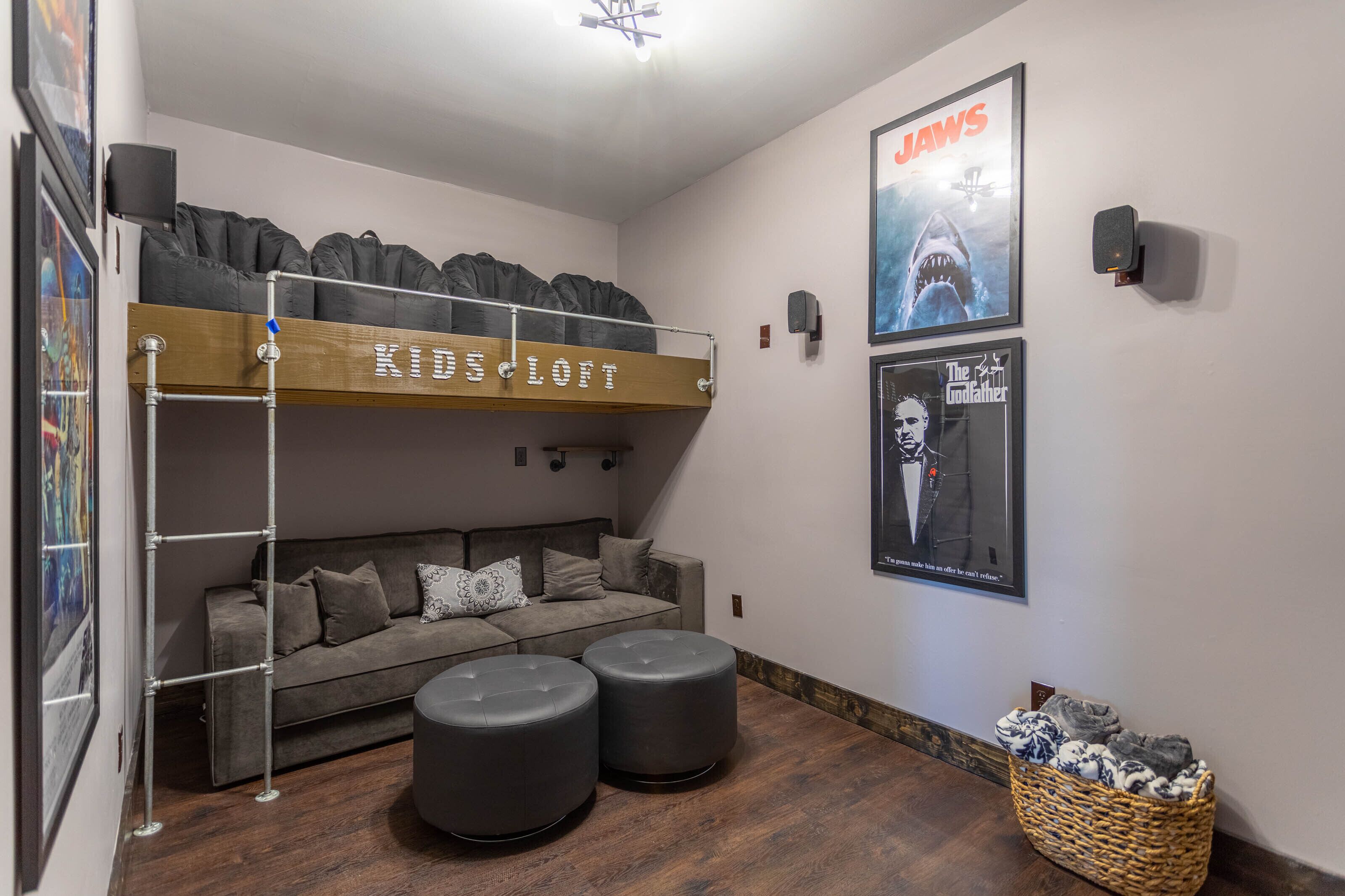 A room with a bunk bed labeled "Kids Loft," a sofa, two ottomans, a framed poster of a shark, and a basket with items.