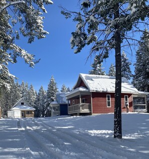 Une cabane recouverte de neige, à l’extérieur rouge, entourée d’arbres chargés de neige et sous un ciel bleu dégagé.