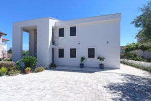 A modern two-story building with a flat roof, white exterior walls, and multiple windows with shutters. There are potted plants and a paved courtyard in front.