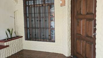 A room with a large wooden door, a window with metal bars, a white textured wall, and a tiled floor.