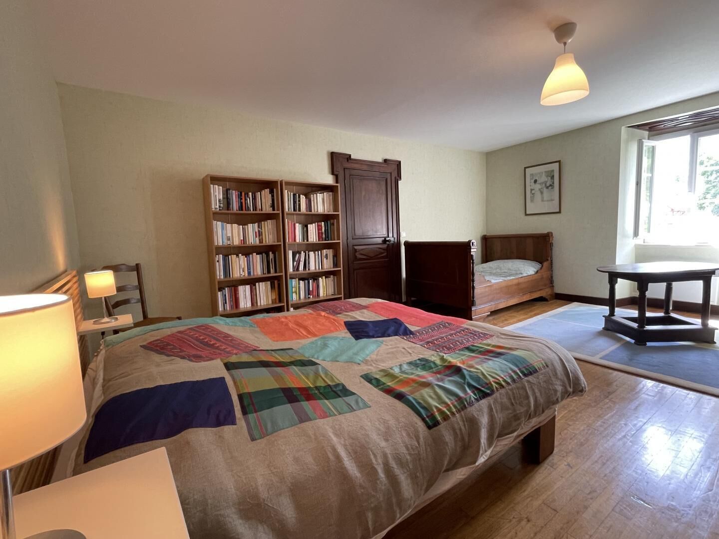 A bedroom with a large bed, a bedside table, a lamp, a chair, a bookshelf, and a window.