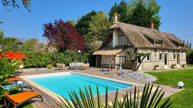 Une maison à toit de chaume avec une piscine, entourée de verdure et d’une pelouse bien entretenue.