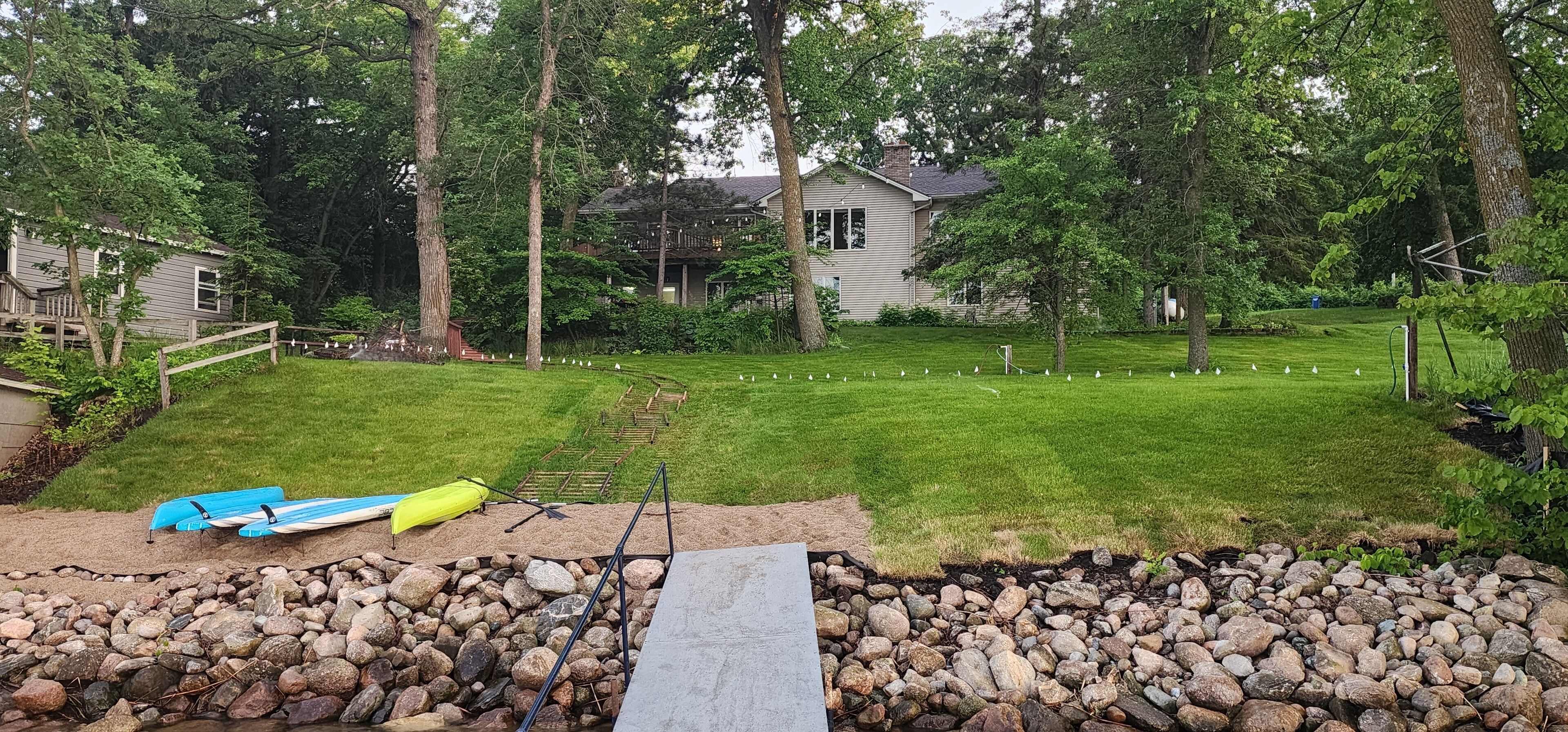 A backyard with a grassy lawn, a wooden deck, and a small pond with a blue kayak and yellow canoe.
