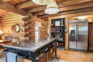 A rustic kitchen with a wooden log wall, a stainless steel refrigerator, and a wooden dining table with chairs. - Doe Creek Ranch | 2BR Cabin with Horse Amenities (Bandera)